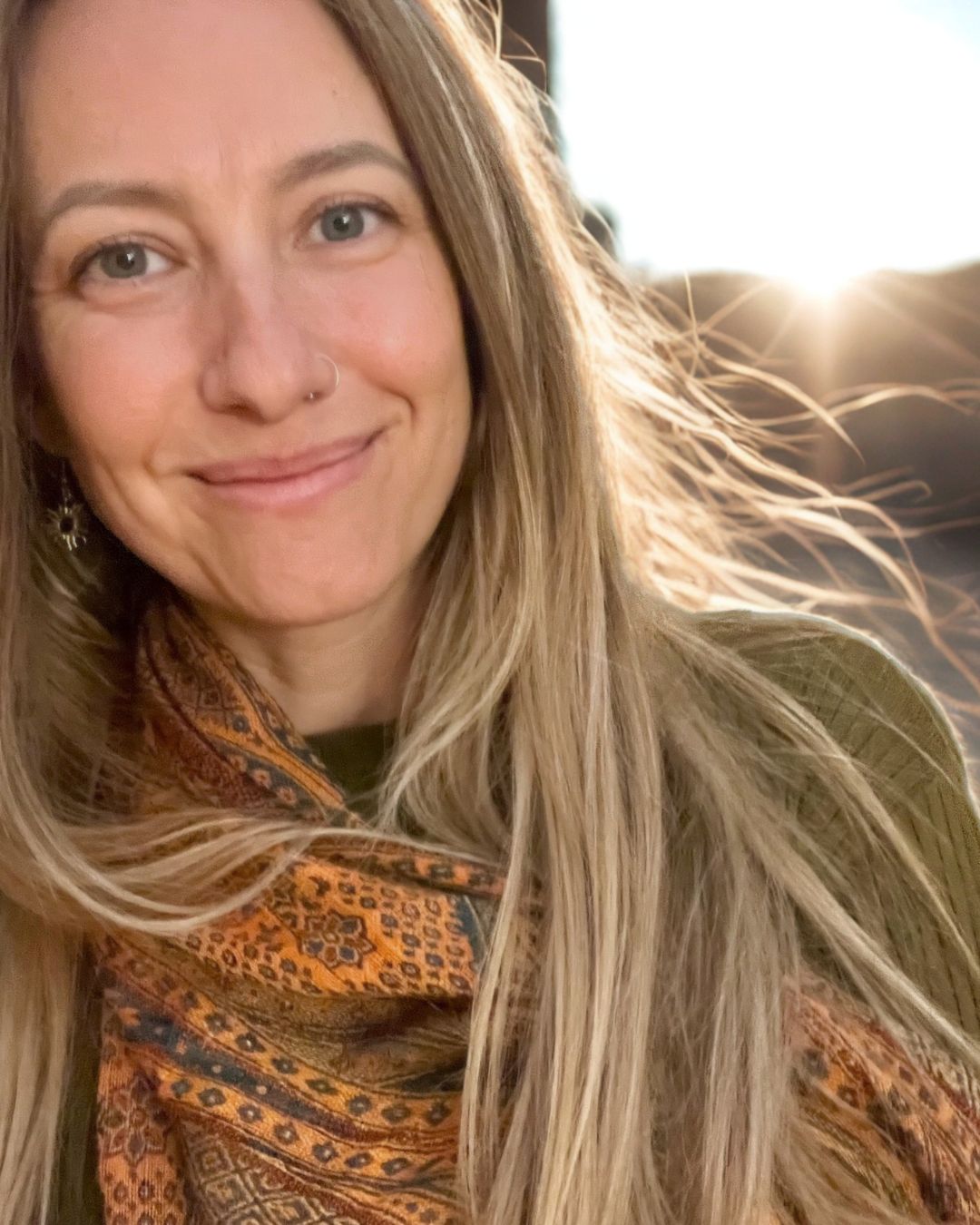 A close-up portrait of a smiling woman with long blonde hair, wearing a patterned orange and brown scarf and a green top, with bright sunlight shining from behind her.